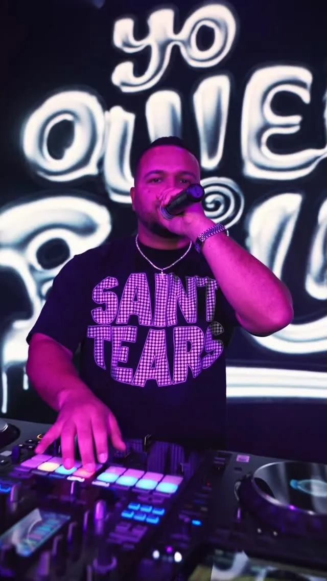 DJ Caises at the CDJs under a Yo Quiero Bailar neon sign, purple wash. Opens larger.